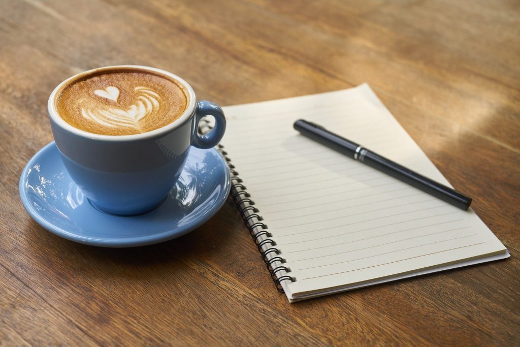 A fancy cup of coffee sitting on a wooden table beside a notebook and pen.