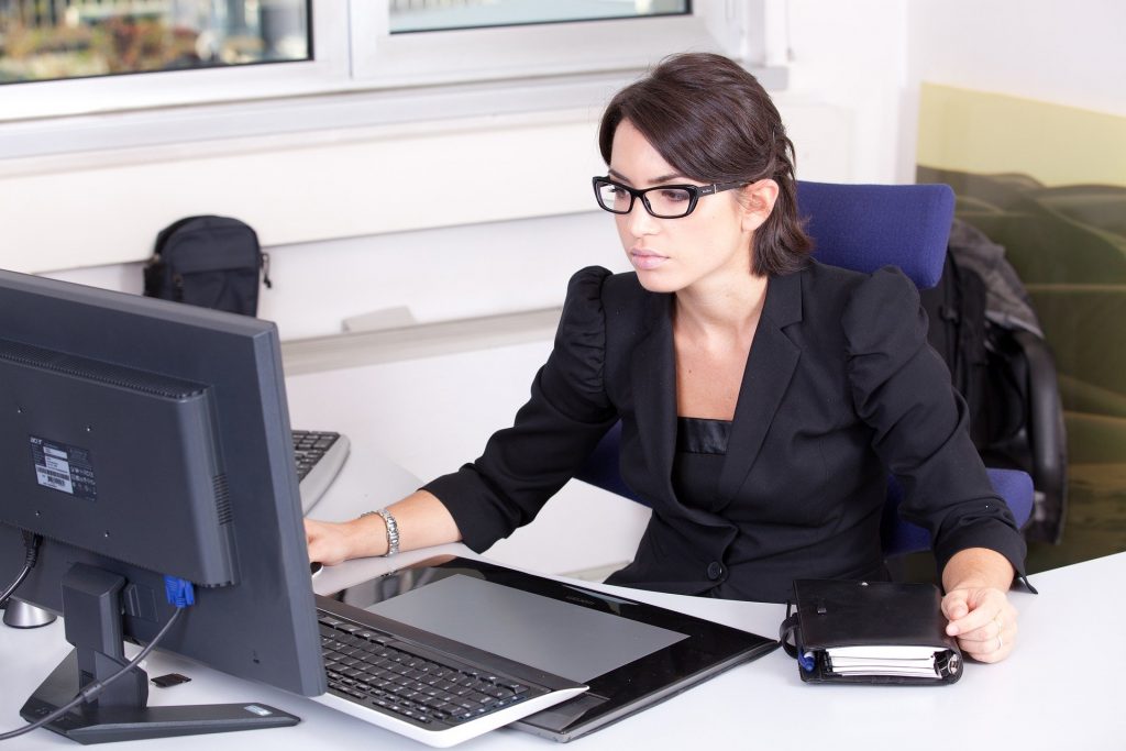 A young woman with brown hair and black glasses sits behind a computer at her office desk.